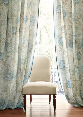 Transparente Gardinen mit einem blassen Blumenmotiv in den Farben Beige, Blau und Taube vor einem eleganten beigen Stuhl.