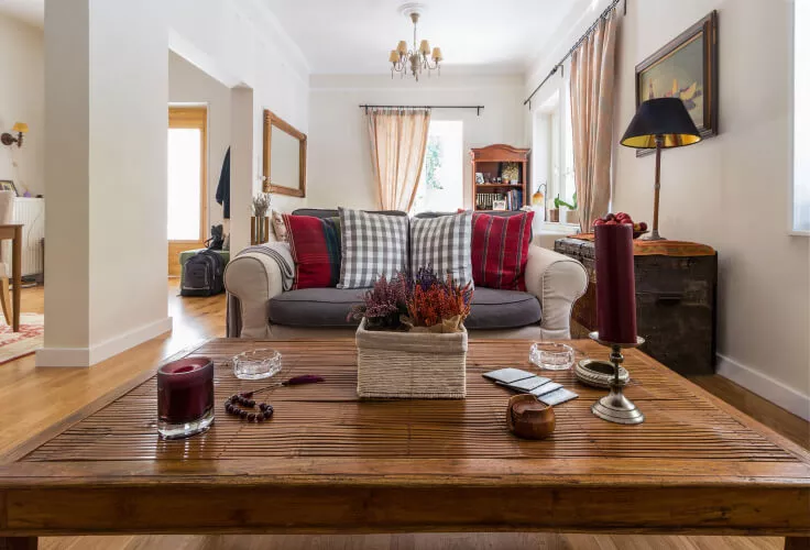 Ein im Landhaus Stil eingerichtetes Wohnzimmer mit orange-grau gemusterten transparenten Vorhängen sowie beiger Couch mit rot gemusterten und karierten Kissen.