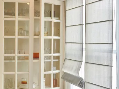 Landhaus-Schrank mit Türen, Gläsern, Teller und orange Windlicht neben Terrassentür mit grauen Faltrollos.