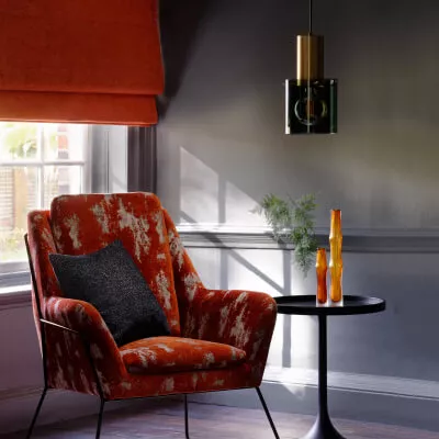 Ein orange-roter Stuhl aus Samtstoff steht in einem dunkel gehaltenen Wohnzimmer mit rotem Raffrollo.