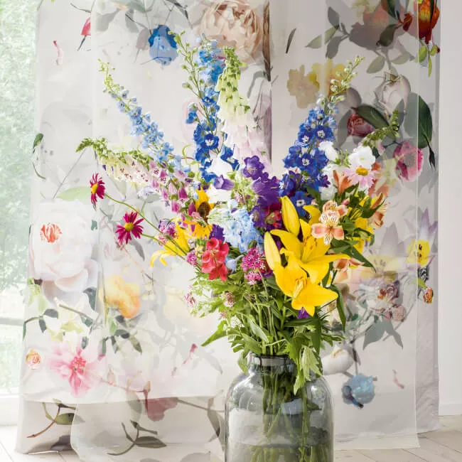 Eindrucksvoll bedruckter, floraler Vorhangstoff, der vor dem Fenster hängt und auf einem hellen Holzboden liegt, hinter einer farbenfrohen Blumenvase