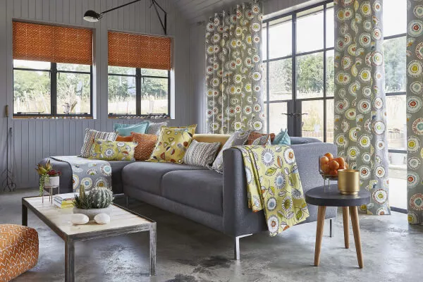 Ein Wohnzimmer im skandinavischen Stil mit Vorhängen und Kissen mit buntem Blumenmuster sowie orangenen Raffrollos.