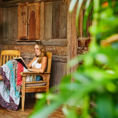 Eine Frau liest ein Buch in einem Stuhl sitzend vor einer gemütlichen antiken Holzfassade.