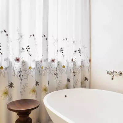 Ein Badezimmer mit weisser Landhausgardine und Pflanzenmotiv hinter einer Badewanne.