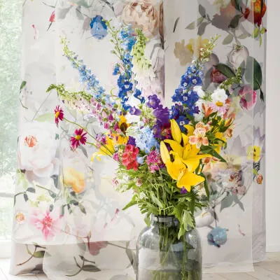 Eine transparente weisse Voile Gardine mit buntem Rosenmotiv hinter einem bunten Blumenstrauß.