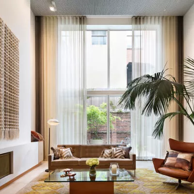 Ein modernes, luxuriöses Wohnzimmer mit Couch und Kissen, sowie transparenten beige-braunen Gardinen vor sehr großen Fenstern.