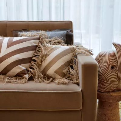 Eine weisse transparente Gardine hinter einem braun-beigem Sofa mit gestreiften Kissen.