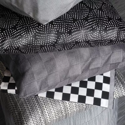 Graue Kissen in verschiedenen Mustern und Texturen, gestapelt auf einem strukturierten grauen Hocker
