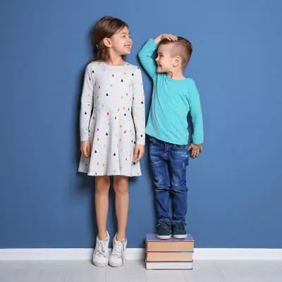 Ein Junge steht vor einer blauen Wand auf Büchern um größer zu sein als seine Schwester, die ihn aber überragt.