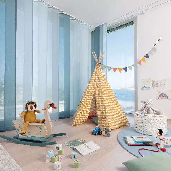 Hell gestaltetes Kinderzimmer mit blauen Flächenvorhängen an einem Eckfenster, gelbem Zelt und und vielfältiges Spielzeug.