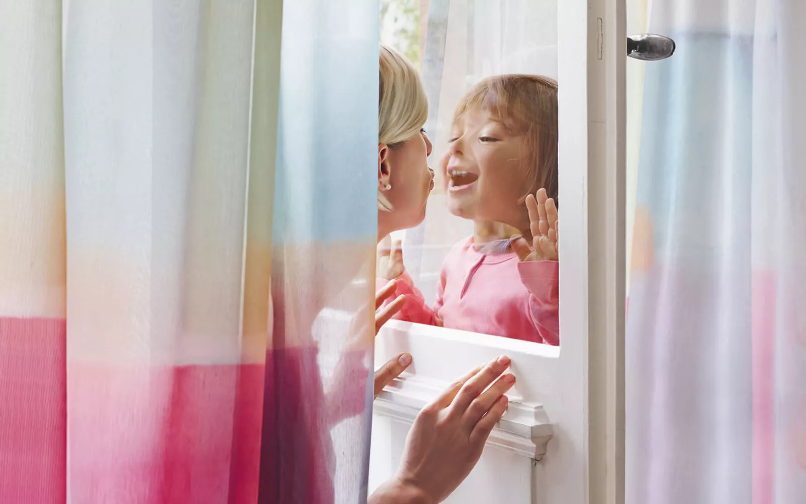 Interaktion zwischen Mutter und Kind durch eine Fensterscheibe, umrahmt von farbigen Gardinen in Weiß, Orange und Pink