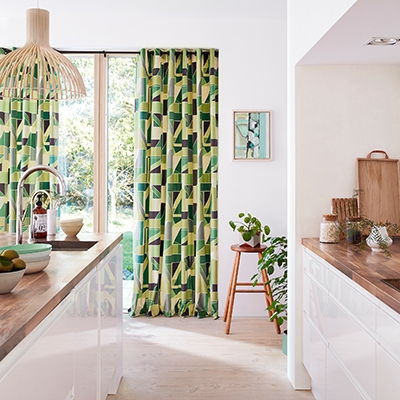 Vorhänge in verschiedenen Grüntönen und Gelb mit einem grafischen Muster vor einem Fenster, durch das man einen Garten sieht, schaffen einen Akzent in einer Küche mit weißem Mobiliar und einer holzfarbenen Arbeitsplatte.