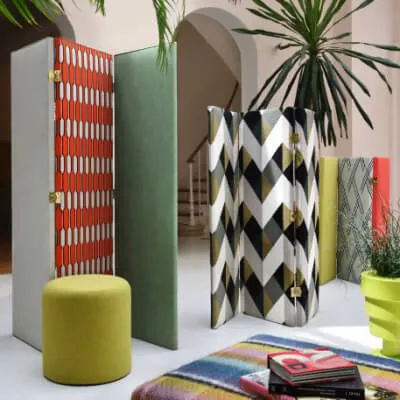 Raum mit cappuccinofarbenen Wänden, Paravents in verschiedenen Mustern & Farben vor Rundbögen mit Holztreppe im Hintergrund.