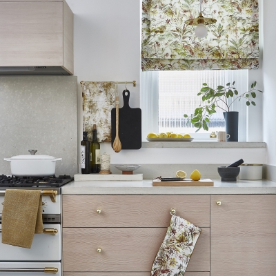 Küche mit farbenfrohem Raffrollo in Fensternische, Küchenutensilien im Vordergrund und Pflanzen auf dem Fensterbrett.