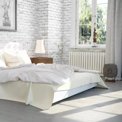 Ein Schlafzimmer im Altbau mit heller Backsteinwand, hellgrauen Vorhängen und weisser Bettwäsche.