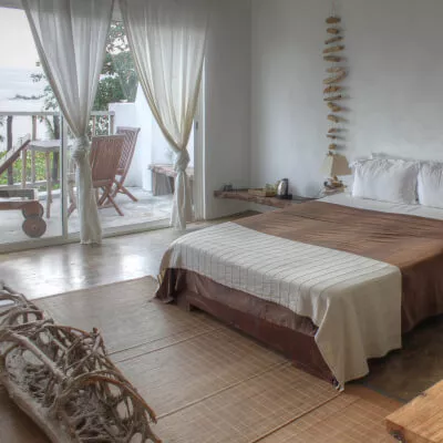 Bodentiefe Fenster mit verknoteten weißen Vorhängen; Blick aufs Meer, Holzmöbel und natürliche Töne dominieren das Schlafzimmer.