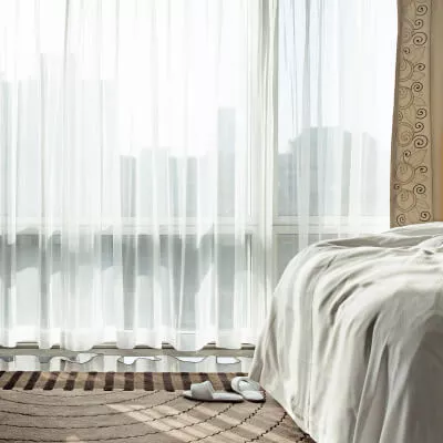 Eine Detailaufnahme von einem Schlafzimmer mit transparent-weissen bodenlangen Gardinen sowie Hausschuhen.