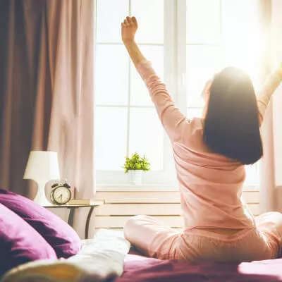 Eine Frau wacht morgens im Schlafzimmer auf und durch die offenen Vorhänge strahlt Sonnenlicht ins Zimmer.