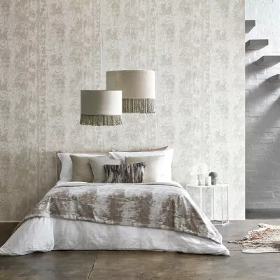 Schlafzimmer mit grau-beige-braun gemusterter Tapete und Bett mit drapierten Decken sowie Leuchten und Tisch